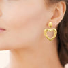 Boucles d'oreille pendantes XL Paloma, puce ovale et pendant cœur ajouré. En acier PVD doré texturé effet tweed. Présentées portées.